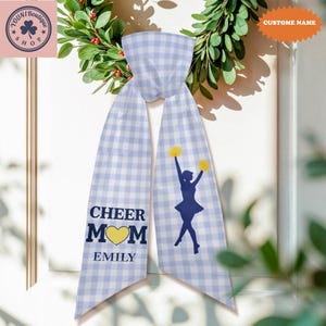 Puede incluir: Una bufanda a cuadros azul claro y blanco con las palabras "CHEER MOM" y un corazón amarillo. También presenta la silueta de una animadora con pompones amarillos. El nombre "EMILY" está impreso debajo de "CHEER MOM".