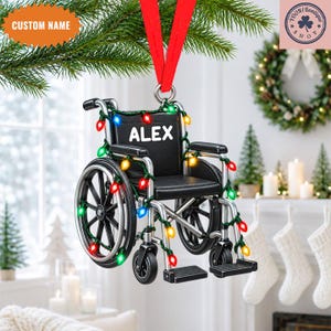 Peut inclure: Une décoration de Noël suspendue en forme de fauteuil roulant noir et argent, ornée d'une guirlande de lumières festives rouges, vertes, bleues et jaunes. Le dossier du fauteuil porte le prénom ALEX imprimé en lettres blanches.