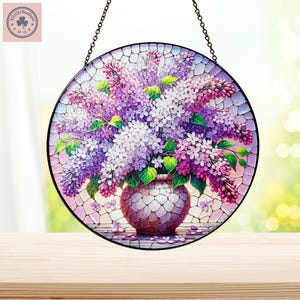 May include: Round stained glass art piece featuring a bouquet of purple and white lilacs in a mosaic vase. The artwork is suspended by a black chain and has a mosaic background. The piece is a decorative item.