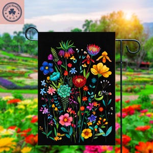 May include: A black garden flag with a colourful floral design. The artwork displays a variety of flowers in shades of blue, red, yellow, and purple, set against a black backdrop. The flag is mounted on a black metal stand.