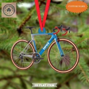 May include: A blue and silver bicycle ornament with red accents, a red ribbon, and the name "SOPHIA" on the frame. The wheels have a glowing orange outline. The ornament is hanging against a blurred green background. The text "CUSTOM NAME" is visible.