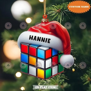 Puede incluir: Adorno navideño con diseño de Cubo de Rubik, coronado con un gorro de Papá Noel rojo. El nombre "HANNIE" está impreso en el gorro. El adorno es un artículo plano 2D, con una cuerda dorada para colgar. El fondo es un árbol de Navidad borroso.
