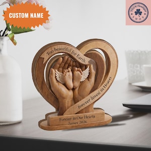 May include: A wooden heart-shaped memorial with the text "Too beautiful for Earth, forever loved in Heaven." The design features hands cradling baby feet with angel wings. The base reads "Forever in Our Hearts James 2026."