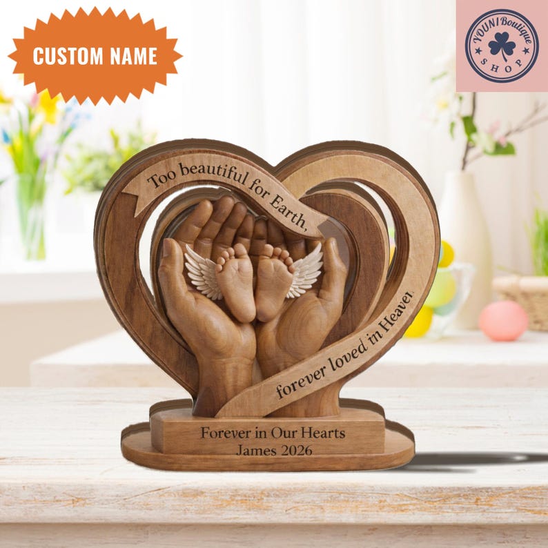 May include: A wooden heart-shaped memorial with the text "Too beautiful for Earth, forever loved in Heaven." The sculpture features hands cradling baby feet with wings. The base reads "Forever in Our Hearts James 2026."