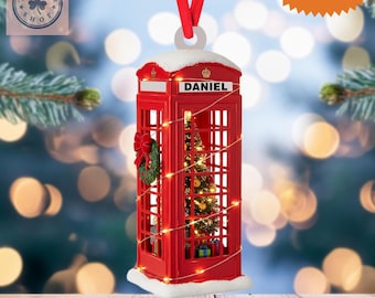 May include: A red telephone booth Christmas ornament with the name "DANIEL" on the front. The ornament has a small Christmas tree inside, a wreath, and string lights. It is a 2D flat item with a red ribbon for hanging.