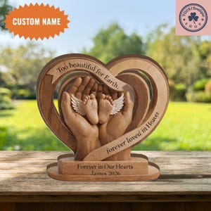 May include: A wooden heart-shaped memorial with the text "Too beautiful for Earth, forever loved in Heaven." The design features hands cradling baby feet with wings. The base reads "Forever in Our Hearts James 2026." The top has space for a custom name.