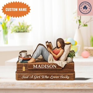 Puede incluir: Figura marrón de una persona leyendo un libro reclinada sobre una pila de libros. La figura tiene una parte superior roja y pantalones grises. El nombre "MADISON" se muestra en los libros. También se ve el texto "Just A Girl Who Loves Books".