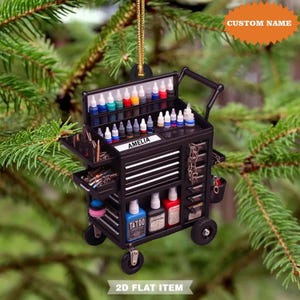 May include: Black miniature tattoo artist's tool chest ornament with wheels, drawers, and a handle. The ornament is filled with colorful ink bottles and tools. The name "Amelia" is printed on a label. The text "CUSTOM NAME" and "2D FLAT ITEM" are also visible.