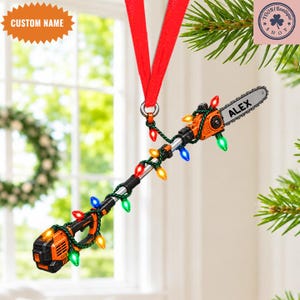 Op de afbeelding: Een oranje en zwarte stokzaag-ornament hangend aan een rood lint. Het gereedschap is omwikkeld met een snoer van kleurrijke kerstverlichting en op het zaagblad staat de naam ALEX in zwarte letters. Het ontwerp bevat een accu aan de onderkant.