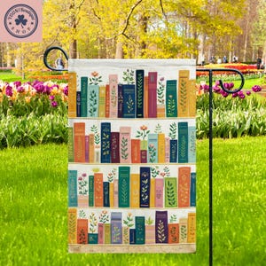 May include: A garden flag featuring a colorful bookshelf design. The flag displays rows of books in various colors, with floral and botanical accents. The flag is displayed outdoors in a garden setting.