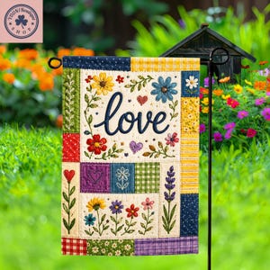 Puede incluir: Una bandera de jardín rectangular con un diseño de patchwork que presenta varios patrones florales, corazones y la palabra love bordada en letra cursiva azul oscuro. La bandera incluye secciones de tela roja, amarilla, verde, azul y morada.