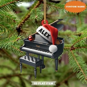 May include: A black piano Christmas ornament with a red Santa hat that says "ALEX". The ornament has a matching piano bench and a gold string for hanging. The background is a blurred image of a Christmas tree.