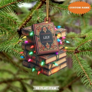 May include: A stack of decorative book ornaments with the name "LILY" on the front. The books are brown with gold accents and are wrapped with colorful Christmas lights. The ornament hangs from a gold string against a backdrop of green pine needles.