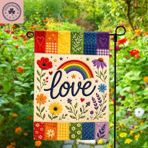 May include: A rectangular garden flag featuring a cream-coloured centre with the word love embroidered in dark blue script. Above the text is a colourful rainbow and stars. The design includes embroidered flowers and hearts, framed by a patchwork border.