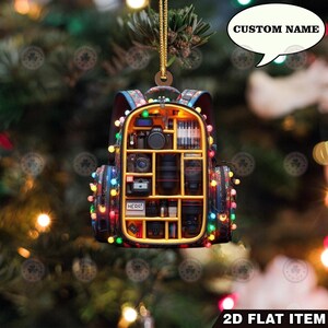 Può includere: Un ornamento per l'albero di Natale a forma di zaino fotografico, pieno di attrezzature fotografiche in miniatura. L'ornamento è decorato con luci colorate e presenta il testo "CUSTOM NAME" in un fumetto. Le parole "2D FLAT ITEM" sono in basso.