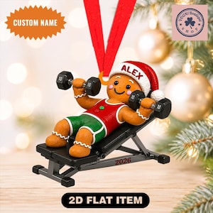 May include: A gingerbread person ornament wearing a red and white Santa hat with the name ALEX printed on it, a red tank top, and green shorts. The figure is lying on a black weight bench lifting two black dumbbells. The year 2026 is printed on the bench.