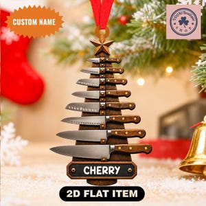 May include: A flat, Christmas tree-shaped wooden ornament featuring seven stacked kitchen knives with brown handles and silver blades. A brown star sits at the top, and a black rectangular plaque at the base displays the name CHERRY in white.