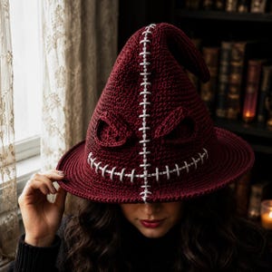 May include: A deep burgundy crocheted hat with a pointed crown and a wide brim. The hat features a prominent vertical seam and a horizontal mouth-like detail, both stitched with thick light gray yarn, creating a textured, expressive face.
