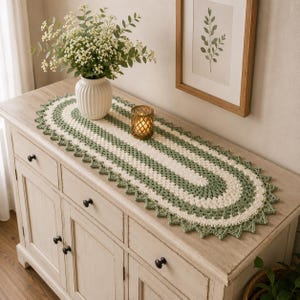 May include: An oval crochet table runner featuring alternating bands of cream and sage green yarn. The runner has a decorative scalloped edge and is displayed on a light wood cabinet with a white vase of flowers and a small candle holder.