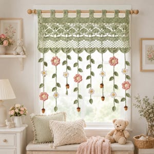 Può includere: Una mantovana all'uncinetto color verde salvia appesa a un bastone di legno, caratterizzata da un bordo superiore smerlato e da molteplici fili verticali pendenti decorati con fiori rosa, fiori bianchi e piccole ghiande marroni.