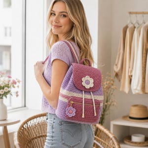 May include: A crochet backpack featuring a purple and cream striped body with a dark pink flap and front pocket. The bag is adorned with a large white crocheted flower on the flap and a smaller purple crocheted flower charm on the gold zipper.