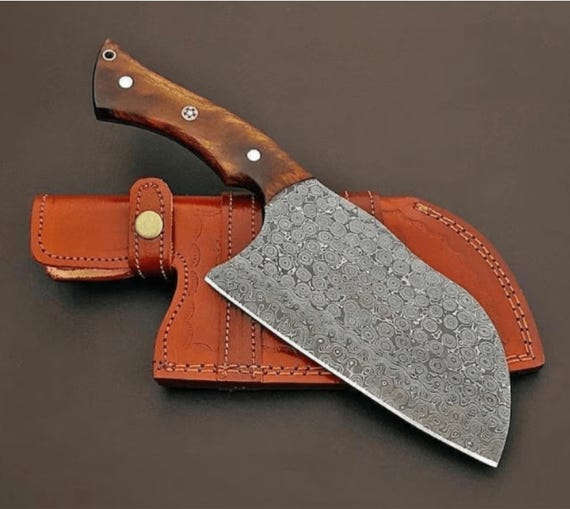 A visually stunning hand-forged Damascus steel cleaver with that unmistakable wavy pattern — the kind of blade that lives on a magnetic strip as kitchen art as much as a serious tool.