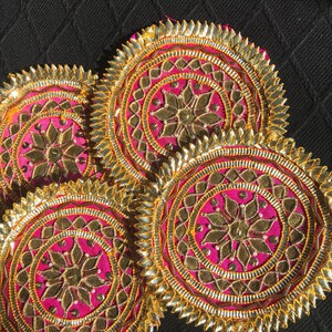 May include: Four decorative, circular coasters with a vibrant pink center, gold embroidery, and a gold, scalloped edge. The coasters feature a floral design in the center and are set against a black background. These coasters add a touch of elegance.