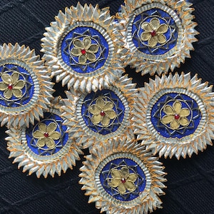 May include: A collection of seven decorative circular embellishments. Each features a central blue circle with a gold flower and red center, surrounded by gold trim and silver, pointed edges. The items are arranged on a black surface.