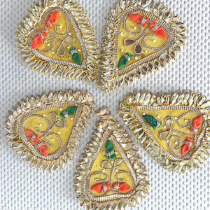 May include: Five yellow, heart-shaped fabric embellishments with gold and silver trim. Each has orange, green, and brown embroidered details. The embellishments are arranged on a white textured surface.