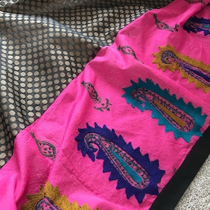May include: A vibrant pink textile with embroidered paisley patterns in blue, teal, and gold. The fabric is layered over a black and gold dotted textile, creating a striking contrast. The edges are trimmed with black.