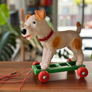 Könnte beinhalten: Ein Vintage-Spielzeug-Hund auf einem grün-roten Wagen. Der Hund ist cremefarben mit orangefarbenen Ohren und Flecken und trägt ein rotes Halsband. Eine rote Schnur ist am Wagen befestigt. Das Spielzeug wird auf einer Holzoberfläche präsentiert.