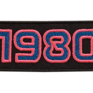 Op de afbeelding: Een zwart rechthoekig embleem geborduurd met het nummer "1980". De cijfers zijn blauw met een roze omtrek. De patch heeft een zwarte rand en een witte achtergrond.