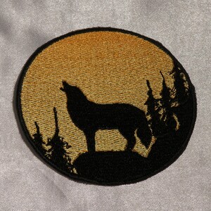 Könnte beinhalten: Gestickter Aufnäher mit einer schwarzen Silhouette eines heulenden Wolfs vor goldenem Hintergrund. Der Wolf steht auf einem schwarzen Hügel mit silhouettierten Bäumen. Der Aufnäher ist rund mit schwarzem Rand.