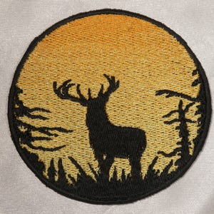May include: Embroidered patch featuring a black silhouette of a deer with large antlers against a golden sunset. The deer stands in a grassy area with trees on either side, all within a black circular border. The background has a gradient of yellow to orange.