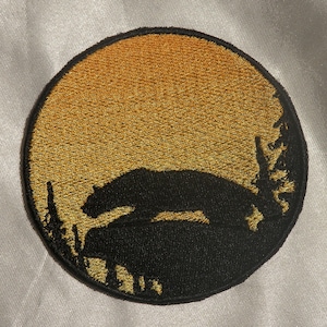 Puede incluir: Parche bordado circular con una silueta negra de oso sobre un fondo de puesta de sol amarillo dorado. El parche tiene un acabado detallado y texturizado, ideal para ropa o accesorios.