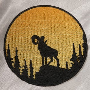 Op de afbeelding: Ronde geborduurde patch met een zwart silhouet van een ram tegen een gouden zonsondergang. Het ontwerp bevat gedetailleerde bomen en een getextureerde, gouden achtergrond, wat een schilderachtig beeld creëert.