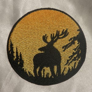May include: Embroidered patch featuring a black silhouette of a deer with large antlers, set against a golden yellow sunset within a circular frame. The design includes trees and grass, creating a nature scene.