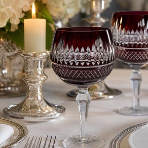 May include: A deep red wine glass featuring a clear crystal stem and base, with intricate diamond and vertical cut patterns etched into the bowl. The glass has a rounded shape and sits on a clear pedestal, showcasing a contrast between the dark red glass and the transparent stem.