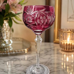 May include: A deep magenta colored crystal wine glass featuring intricate cut glass patterns including starbursts and grape clusters. The stem is clear and faceted, leading down to a circular base with a radial sunburst cut design.