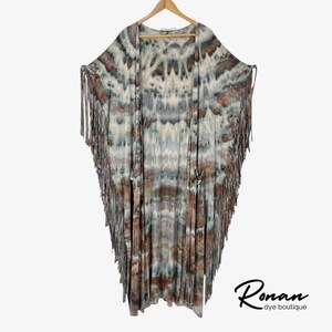 May include: A tie-dye kimono-style robe with fringe trim, hanging on a wooden hanger. The robe features a pattern of white, brown, and blue hues. The bottom of the robe has long fringe. The words "Ronan dye boutique" are in the bottom right corner.