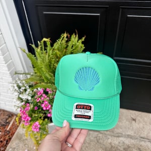 May include: A bright green trucker hat featuring a mesh back and a foam front panel. The front displays an embroidered blue seashell design. A rectangular sticker on the brim reads OTTO WORLD FAMOUS FOAM FRONT OSFM TRUCKER DURABLE SNAP CLOSURE.