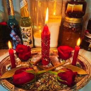 May include: A spiritual arrangement featuring lit red candles, red roses, and glass jars. The candles are placed on a silver tray with herbs and spices. Bottles with labels are in the background, creating a mystical ambiance.