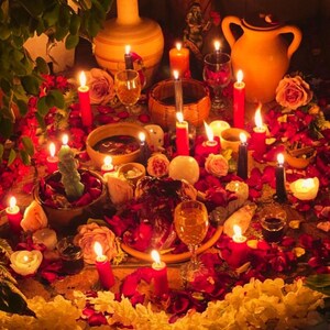 May include: A spiritual altar with lit candles, flowers, and various ritual objects. Red and black candles are interspersed among rose petals and other floral arrangements. Several glasses and ceramic vessels are also present, creating a warm, inviting atmosphere.