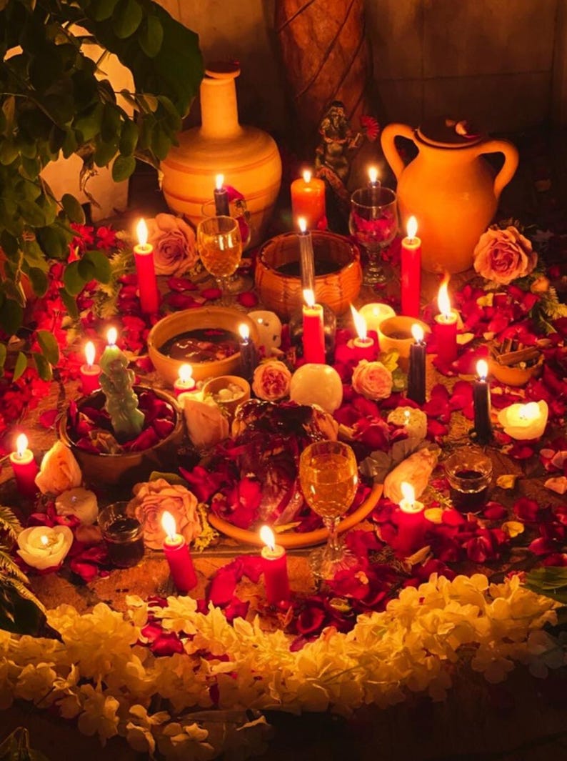 May include: A spiritual altar with lit candles, flowers, and various ritual objects. Red and black candles are interspersed among rose petals and other floral arrangements. Several glasses and ceramic vessels are also present, creating a warm, inviting atmosphere.