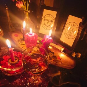May include: A dimly lit scene featuring lit candles, a glass of wine with flower petals, and framed illustrations. A small Eiffel Tower replica and a vase of flowers are also visible. The overall aesthetic suggests a mystical or spiritual theme.