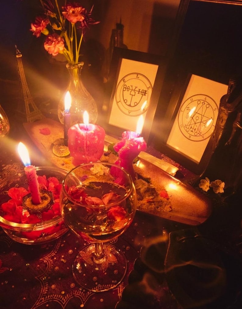 May include: A dimly lit scene featuring lit candles, a glass of wine with flower petals, and framed illustrations. A small Eiffel Tower replica and a vase of flowers are also visible. The overall aesthetic suggests a mystical or spiritual theme.