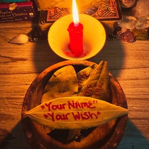 May include: A lit red candle in a yellow bowl sits above a wooden bowl with bay leaves. One leaf has the text "Your NAME" and "Your Wish" in red. Other items include a tarot deck and a bottle.