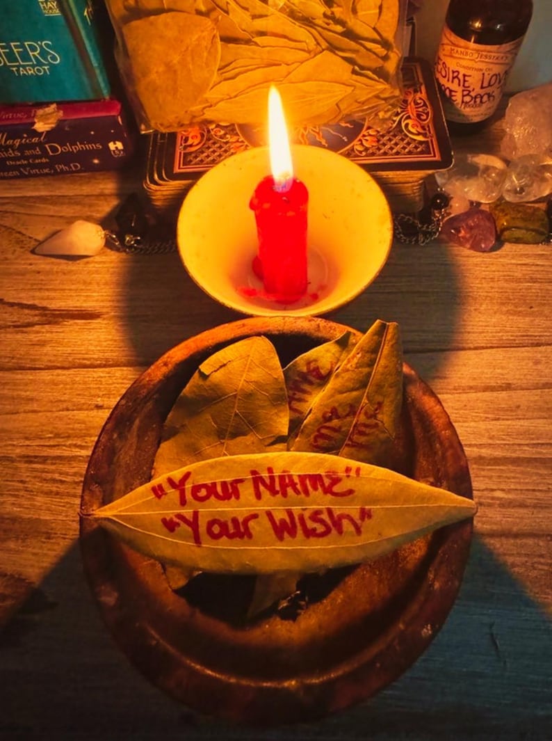 May include: A lit red candle in a yellow bowl sits above a wooden bowl with bay leaves. One leaf has the text "Your NAME" and "Your Wish" in red. Other items include a tarot deck and a bottle.
