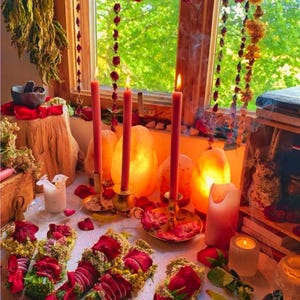 May include: A mystical scene featuring lit red candles in brass holders, surrounded by rose petals and bundles of herbs. Dried roses and greenery hang from the window, creating a warm, inviting atmosphere. The scene evokes a sense of ritual and natural beauty.
