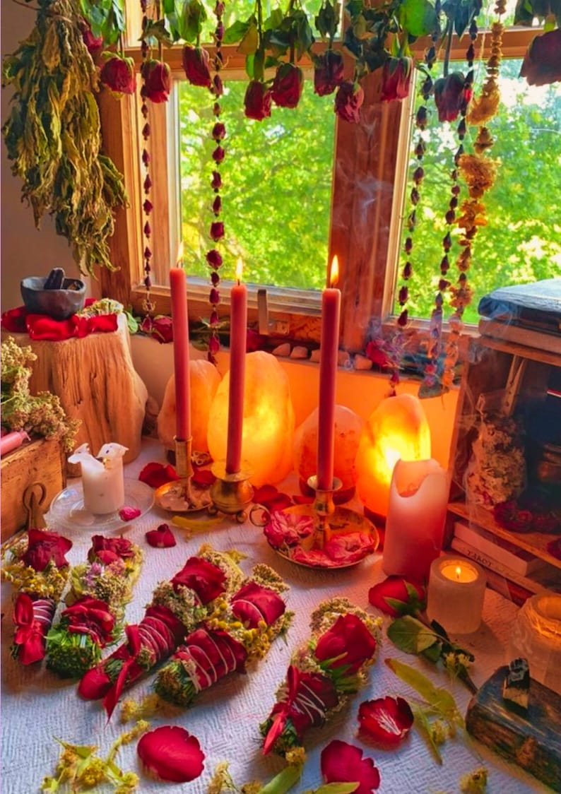 May include: A mystical scene featuring lit red candles in brass holders, surrounded by rose petals and bundles of herbs. Dried roses and greenery hang from the window, creating a warm, inviting atmosphere. The scene evokes a sense of ritual and natural beauty.